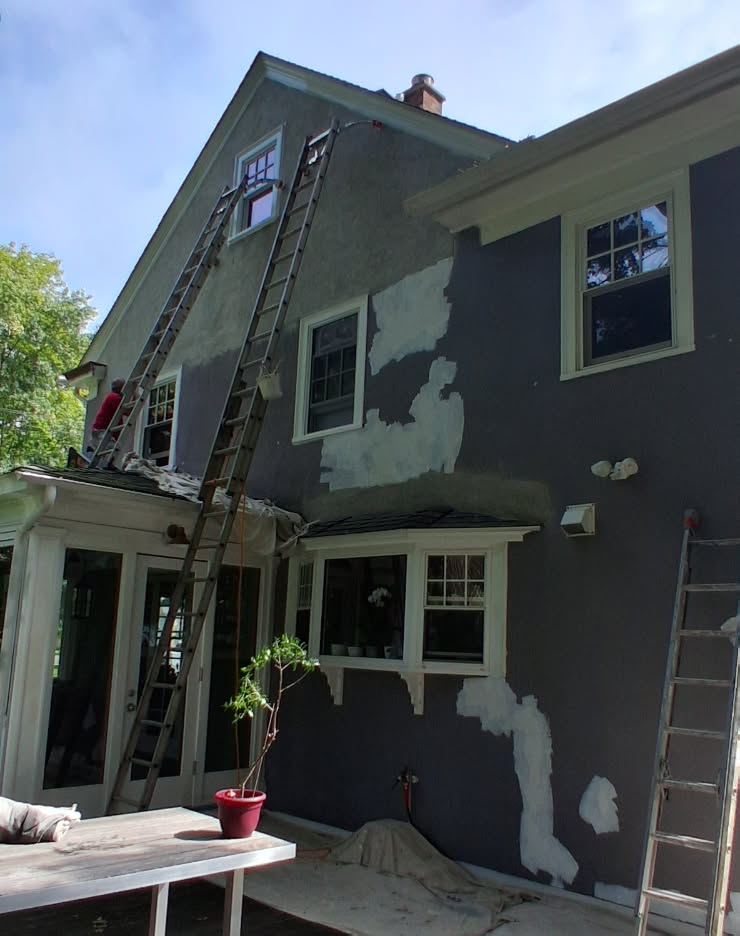 Stucco colonial mid-prep, with grey siding, patched repairs and ladders set against the gable
